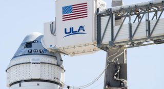 a grey-and-blue conical spacecraft sits atop a white rocket. a metal gantry extends to a hatch on the spacecraft