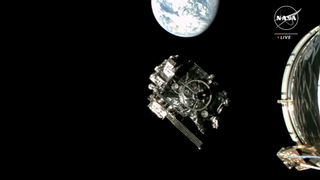 a cube-shaped spacecraft can be seen against the blackness of space. a bright blue-and-white earth can be seen in the background