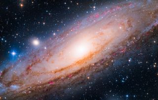 andromeda galaxy bathed in pink. The large spiral galaxy glows orange and pink and is filled with stars, gas and dust. It sits against a backdrop of stars and the blackness of space.